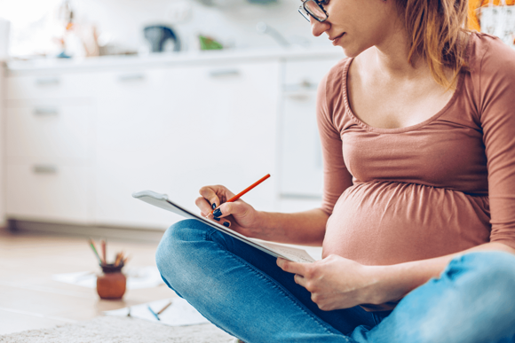 A pregnant woman taking notes about her first trimester while sitting on the floor.